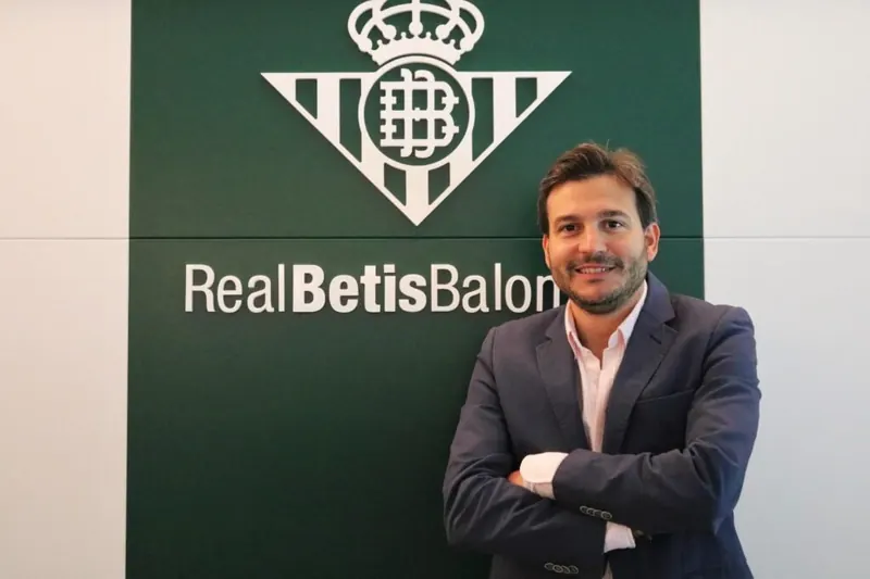 Miguel Roda, con traje, delante de un panel del Real Betis Balompié